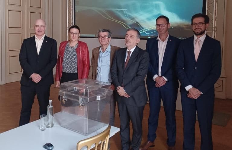 les conseillers consulaires, l'ambassadeur et la chef de section consulaire prennent la pose à devant une urne de vote vide